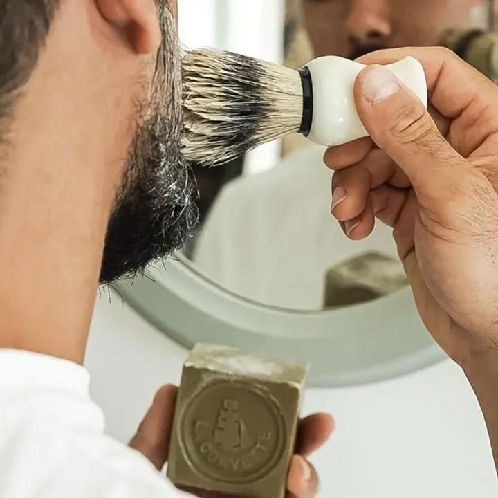 オリーブ石けんを片手に持ち、シェービングブラシで頬に泡を塗っている使用例
